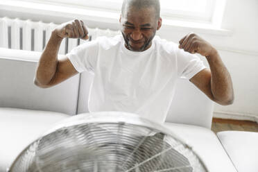 Man flexing muscles in front of electric fan at home - EYAF02618