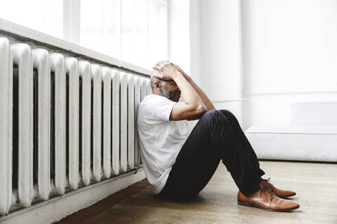 Depressed man with head in hands sitting near heater at home - EYAF02628