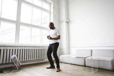 Happy man dancing in front of electric fan at home - EYAF02629