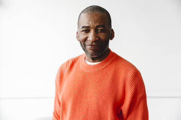 Älterer Mann mit orangefarbenem Pullover vor weißer Wand - EYAF02638
