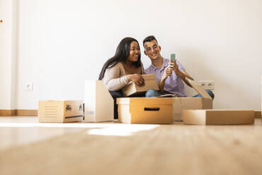 Happy couple taking selfie sitting by cardboard boxes in new home - JCCMF10324