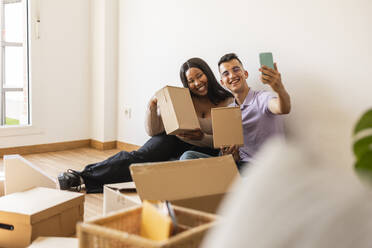 Happy couple taking selfie with cardboard boxes in new home - JCCMF10325