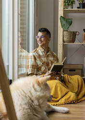Smiling man with book looking out of window at home - JCCMF10379