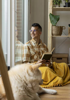 Lächelnder Mann mit Buch, der zu Hause aus dem Fenster schaut - JCCMF10379