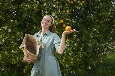 Smiling woman standing near orange tree in orchard - ANNF00149