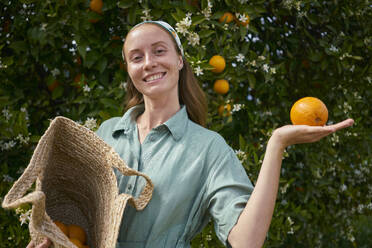 Lächelnde junge Frau mit einer Orange in der Hand in der Nähe eines Baumes im Obstgarten - ANNF00150