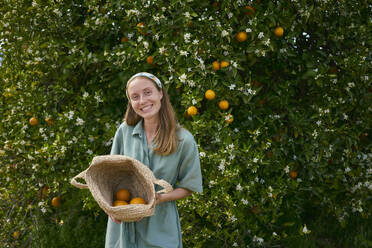 Smiling woman standing with basket of oranges in front of tree at orchard - ANNF00156