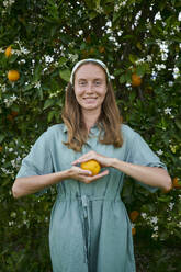 Smiling woman with fresh orange in front of tree at orchard - ANNF00168