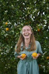 Happy woman with brown hair holding oranges in front of tree at orchard - ANNF00170