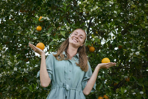 Smiling woman holding oranges near tree in orchard - ANNF00171