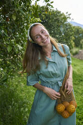 Lächelnde Frau mit Orangen im Netzbeutel auf einer Obstplantage - ANNF00173