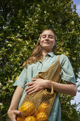 Smiling woman with fresh oranges in mesh bag near tree at orchard - ANNF00178