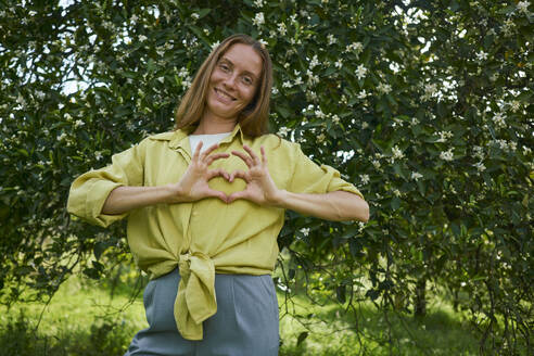 Woman making heart gesture standing in front of tree at garden - ANNF00208