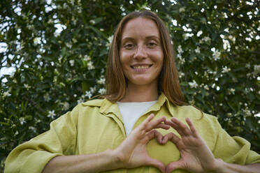 Smiling woman making heart gesture in garden - ANNF00210