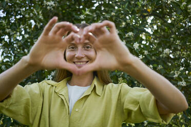 Smiling young woman making heart sign in garden - ANNF00211
