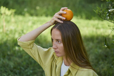 Ernste Frau balanciert Orange auf dem Kopf im Garten - ANNF00213