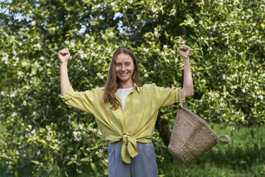 Happy woman carrying straw bag standing in front of tree at orchard - ANNF00220