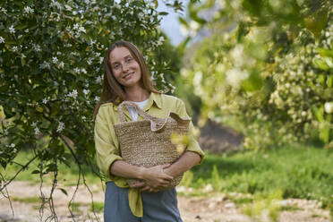 Smiling woman holding straw bag standing in orchard - ANNF00221