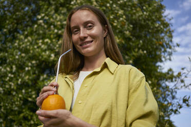 Lächelnde Frau mit Orange und wiederverwendbarem Metallstrohhalm im Obstgarten - ANNF00233