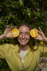 Smiling woman holding slices of fresh oranges in orchard - ANNF00236