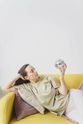 Woman holding glass ball lying on yellow sofa - EYAF02653