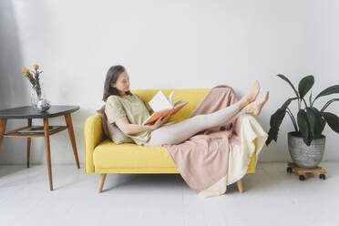 Woman reading book on yellow sofa in living room - EYAF02654