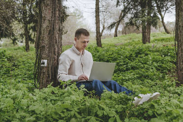Geschäftsmann mit Laptop, der mit einer Steckdose an einem Baum im Wald aufgeladen wird - YTF00777