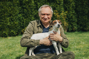 Senior man holding dog sitting on grass in back yard - VSNF00785