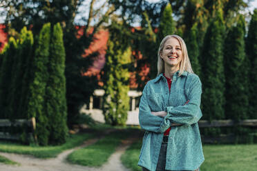 Blond woman standing with arms crossed in front of house - VSNF00787