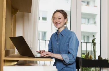 Smiling freelancer wearing denim shirt working on laptop at home - MIKF00247