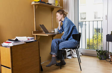 Freelancer in denim shirt looking at laptop and working from home - MIKF00251