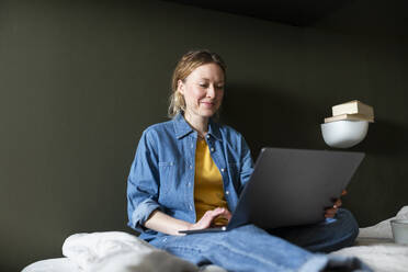 Lächelnder Freiberufler in Jeanshemd mit Laptop auf dem Bett sitzend zu Hause - MIKF00254