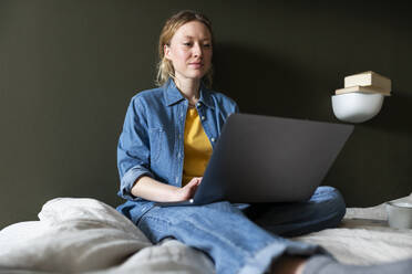 Young freelancer in denim shirt working with laptop sitting on bed at home - MIKF00255