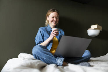 Junger Freiberufler hält Kaffeetasse und arbeitet mit Laptop auf dem Bett - MIKF00256