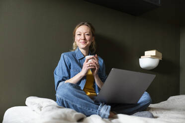 Freelancer hält Kaffeetasse mit Laptop auf dem Bett - MIKF00258