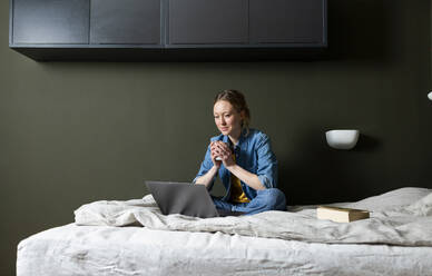 Geschäftsfrau hält Kaffeetasse und arbeitet mit Laptop auf dem Bett - MIKF00262