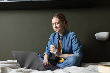 Freelancer mit Kaffeetasse und Laptop auf dem Bett - MIKF00263