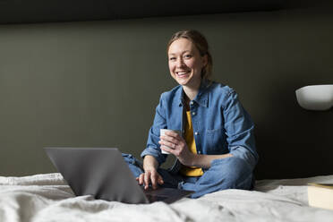 Smiling businesswoman holding coffee cup with laptop on bed - MIKF00264