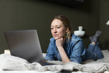 Geschäftsfrau in Jeanshemd arbeitet mit Laptop auf dem Bett liegend - MIKF00267