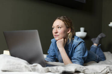 Thoughtful freelancer in denim shirt lying on bed with laptop - MIKF00268