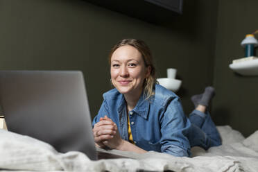 Lächelnder Freiberufler in Jeanshemd mit Laptop auf dem Bett liegend - MIKF00269