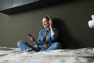 Junge Frau genießt Musik mit Kopfhörern auf dem Bett sitzend - MIKF00270