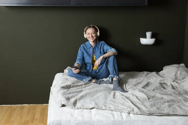 Smiling young woman enjoying music with headphones on bed - MIKF00273
