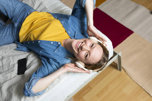 Happy woman enjoying music with headphones lying on bed - MIKF00280