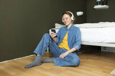 Smiling young woman enjoying music with headphones sitting on floor - MIKF00281