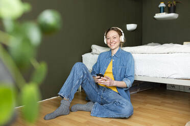 Smiling young woman listening music with headphones sitting on floor - MIKF00282