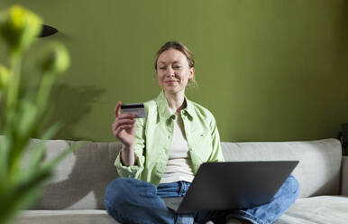 Frau beim Homeshopping mit Laptop und Kreditkarte auf der Couch - MIKF00294