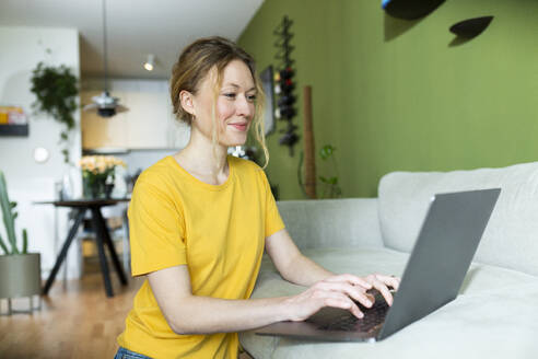 Freiberufler mit gelbem T-Shirt arbeitet mit Laptop auf der Couch - MIKF00296