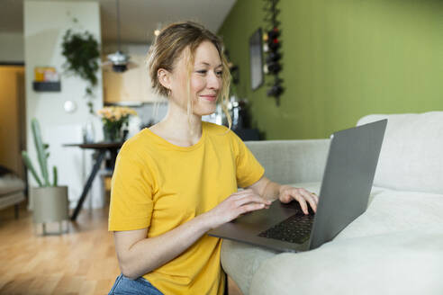 Lächelnder junger Freiberufler in gelbem T-Shirt bei der Arbeit mit Laptop - MIKF00297