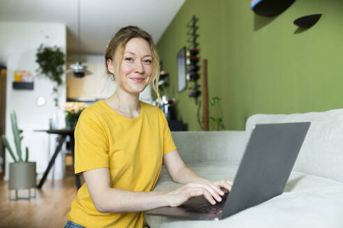 Junger Freiberufler in gelbem T-Shirt bei der Arbeit mit Laptop zu Hause - MIKF00298